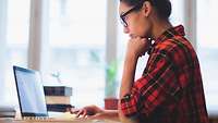 Frau mit Brille am Laptop Dunkelhaarige Frau mit Brille und Flanellhemnd sitzt am Schreibtisch und arbeitet an einem Laptop