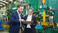 Zwei Mitarbeitende arbeiten am Tablet, im Hintergrund ist eine industriellen Fertigungsanlage