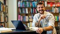 Junger Mann am Schreibtisch mit Büchern lächelt in die Kamera