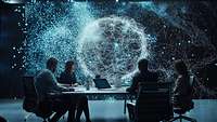 Vier Personen sitzen an einem Tisch mit aufgeklappten Laptops, im Hintergrund ist eine mit Netz umspannte Kugel