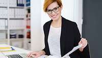 Junge Frau mit Blazer am Schreibtisch tippt in Laptop und blättert in Ordner, im Hintergrund Regale