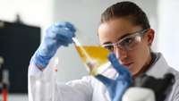 Frau im Laborkittel und mit Schutzhandschuhen blickt in ein Reagenzglasmit einer gelben Flüssigkeit