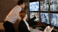 Ein Mann und eine Frau checken Überwachungsvideos auf einer Reihe von Monitoren