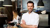 Mann in weißem Hemd und verschränkten Armen steht in einem Restaurant, im Hintergrund Speisen und eine Mitarbeiterin