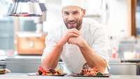 Mann mit Kochmütze stützt sich auf der Durchreiche einer Küche auf, im Vordergrund zwei exklusiv arrangierte Gerichte