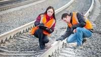 Eine Frau und ein Mann mit Warnwesten checken und protokollieren den Zustand einer Eisenbahnschiene