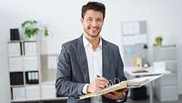 Mann steht im Büro mit geöffneten Ordner und Stift in der Hand