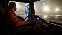 Fahrer mit oranger Jacke sitzt am Steuer eines LKW und steuert nachts über ein großes Betriebsgelände